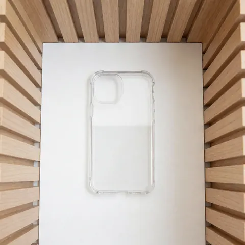 Coque de protection transparente pour smartphone avec découpes précises, posée sur une surface blanche entourée de panneaux en bois clair sous un éclairage diffus et propre.