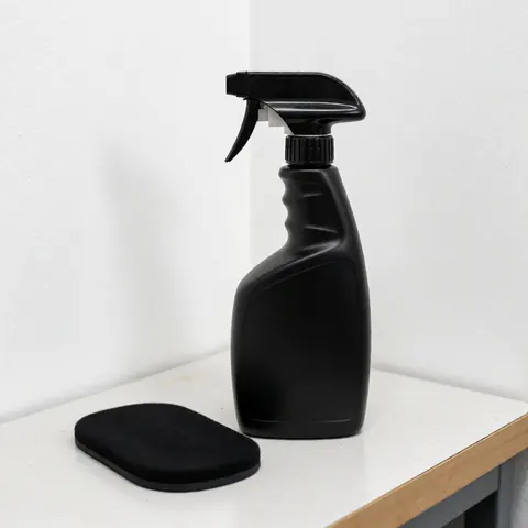 Seau noir avec pulvérisateur et chiffon noir posés sur un banc blanc, murs blancs et éclairage professionnel pour un service de lavage automobile.