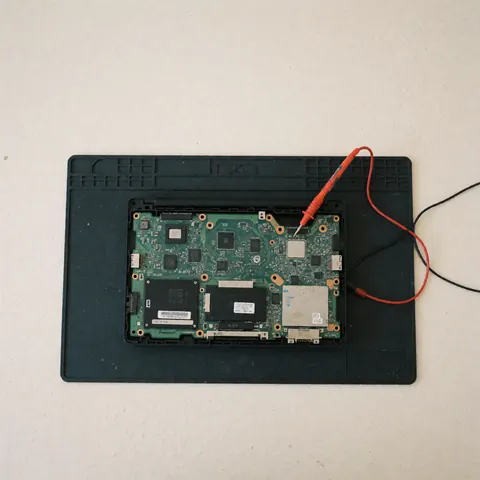 Carte mère d'ordinateur posée sur une surface antistatique avec un multimètre pour diagnostic technique, éclairage uniforme sur fond blanc.