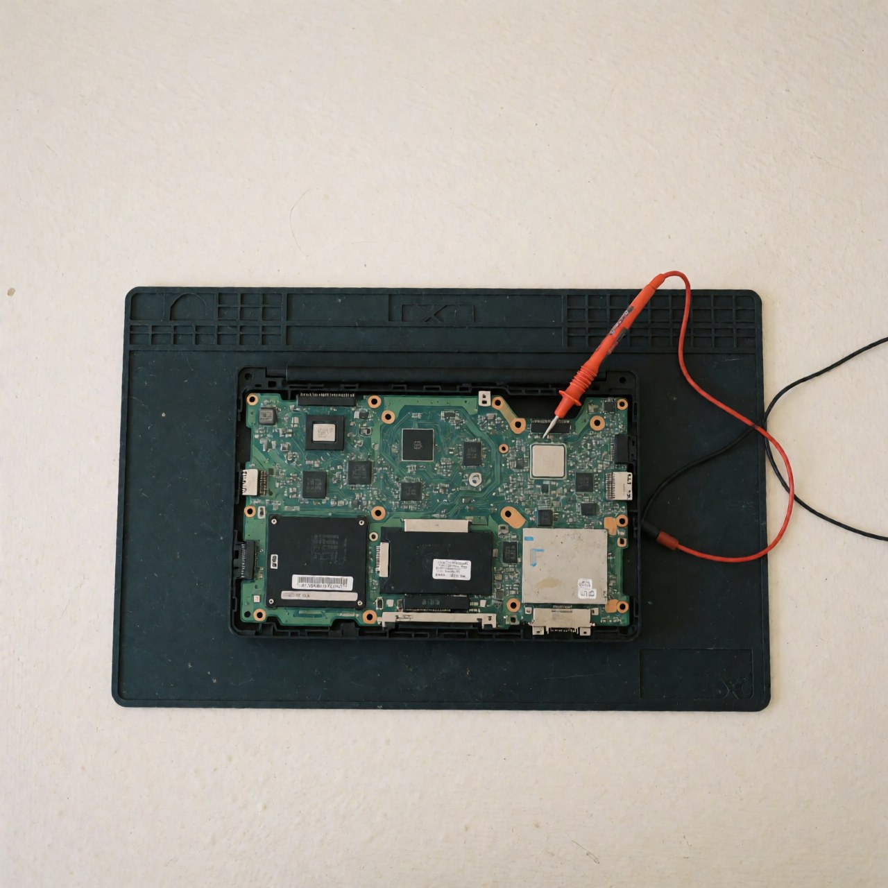 Carte mère d'ordinateur posée sur une surface antistatique avec un multimètre pour diagnostic technique, éclairage uniforme sur fond blanc.