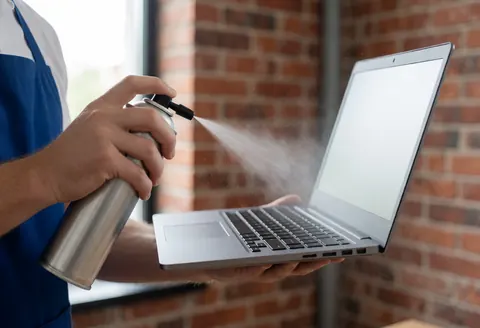 Un technicien en tenue de travail utilise de l'air comprimé pour nettoyer la ventilation d'un ordinateur portable, avec des particules de poussière visibles devant un mur de briques rouges.