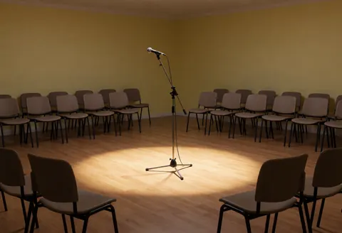 Scène de récital vide avec microphone central, chaises pour le public et éclairage chaud sur le sol en bois.