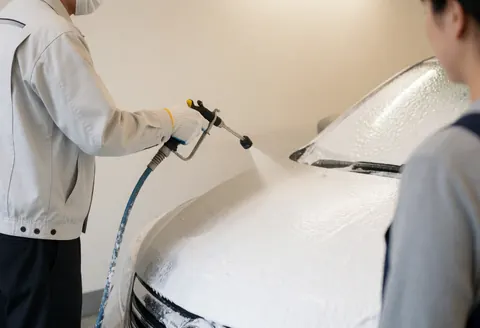 Un employé utilise un tuyau spécial pour projeter de la mousse blanche sur le toit d'un véhicule. Le mur en arrière-plan est d'un blanc chaud. La scène illustre le processus de lavage automobile en cours.