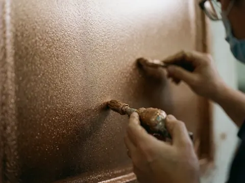 Des mains utilisent un pinceau pour appliquer une peinture métallisée brillante sur une surface, révélant une texture détaillée et des reflets lumineux sur un fond flou.