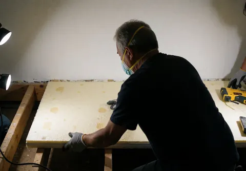 Un professionnel pose un panneau d'isolation thermique sur un plafond de maison, avec des outils, du bois et des lumières visibles.