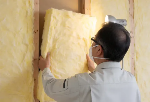 Un ouvrier équipé d'un masque et de lunettes pose un isolant jaune entre des poutres, illustrant les travaux de construction et la sécurité.