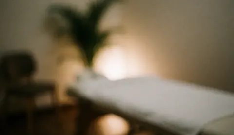 Fond de salle de massage avec lit couvert et plante, éclairage chaleureux et tons terre, idéal pour du texte.