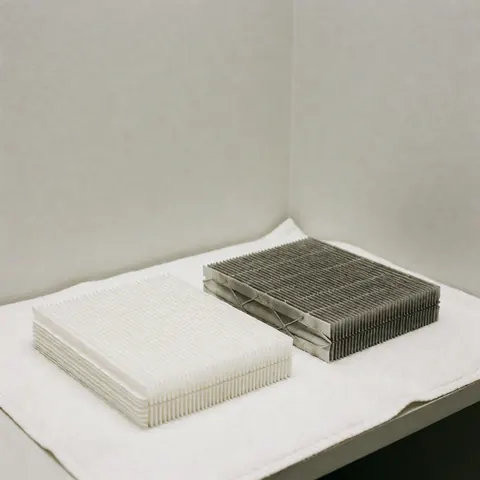 Un filtre de chauffage blanc neuf et un filtre sale usé posés sur un tissu blanc dans un coin de mur gris clair pour illustrer le besoin de remplacement.
