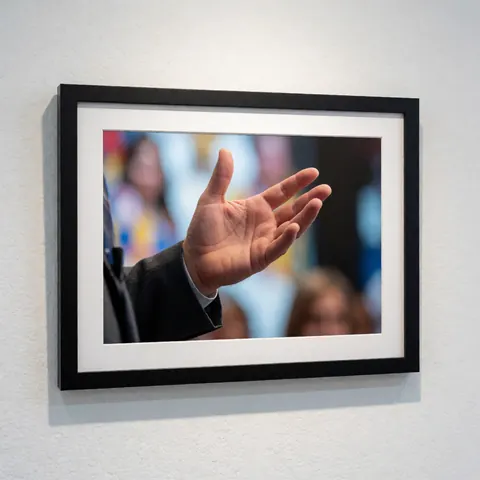 Photographie d'une main effectuant un geste artistique, présentée dans un cadre noir sur un mur blanc texturé avec un arrière-plan flou de personnes.