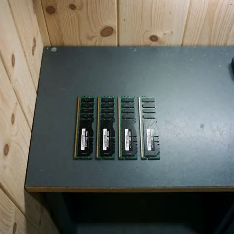 Quatre modules de mémoire RAM DDR4 munis d'étiquettes blanches, disposés en ligne sur une table de travail sombre avec un fond en bois clair.