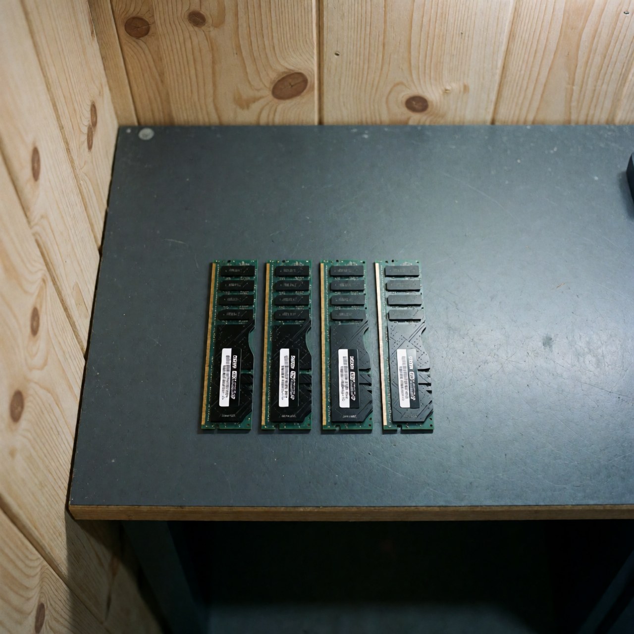 Quatre modules de mémoire RAM DDR4 munis d'étiquettes blanches, disposés en ligne sur une table de travail sombre avec un fond en bois clair.