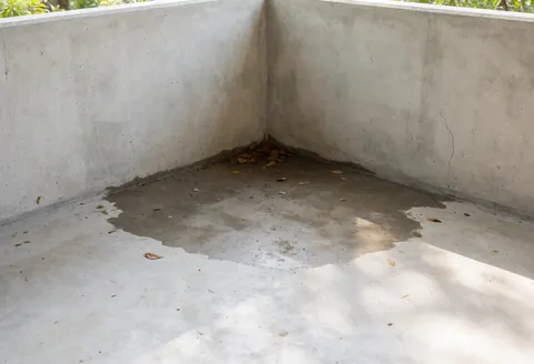 Vue d'un patio en béton avec une tache d'humidité et des feuilles mortes dans un coin, éclairé par la lumière naturelle qui contraste les zones sèches et mouillées.