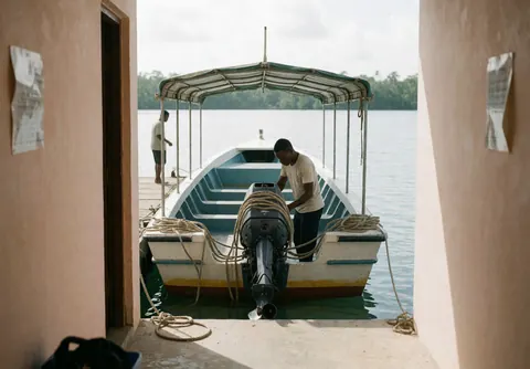 Un employé répare le moteur d'un bateau amarré au quai avec une bâche et des cordes, l'eau calme reflète le ciel et la végétation tropicale en arrière-plan.