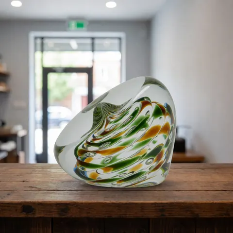 Poids en verre artisanal orné de motifs verts et jaunes posé sur un comptoir en bois rustique dans un atelier de soufflage de verre.