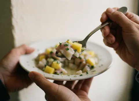 Des mains tenant une cuillère remplie de ceviche frais au poisson, mangue et coriandre sur un fond clair.