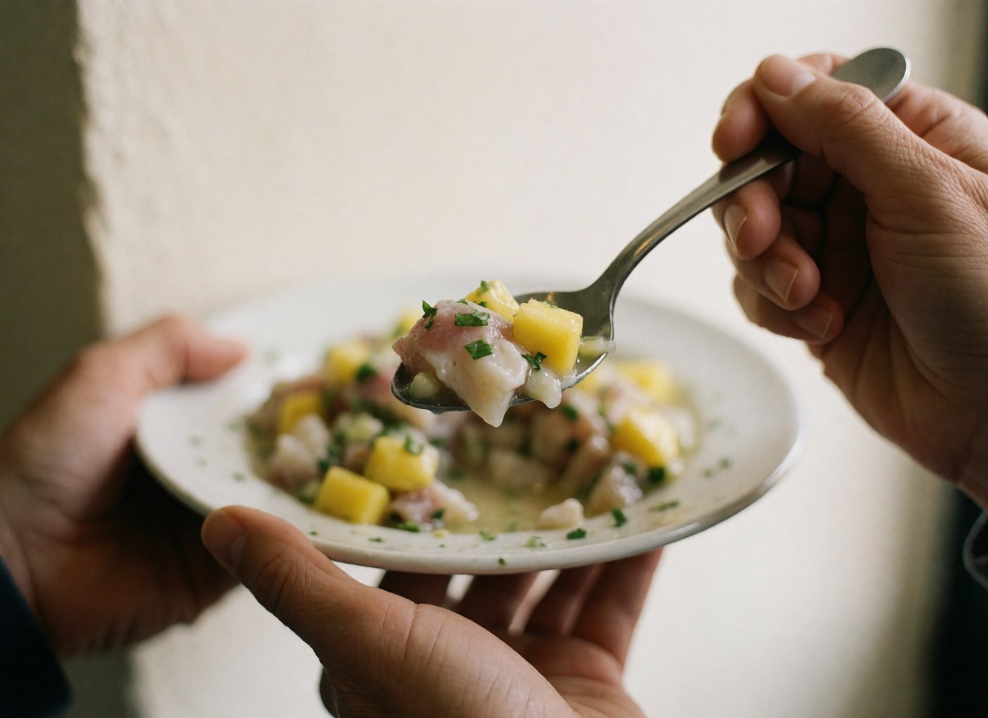 Des mains tenant une cuillère remplie de ceviche frais au poisson, mangue et coriandre sur un fond clair.
