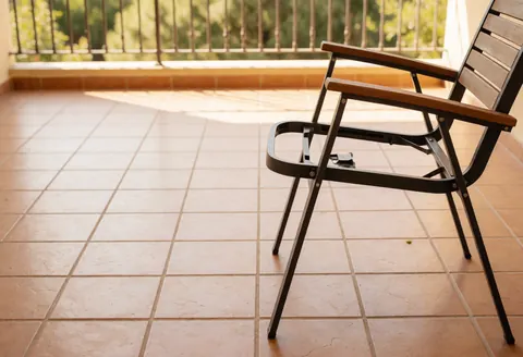 Chaise de jardin métallique avec assise en bois posée sur un sol carrelé, éclairage naturel et rambarde en arrière-plan.