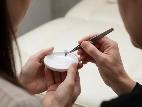 Des pinces professionnelles maintiennent des extensions de cils sur une palette blanche dans un salon de beauté au fond flou.