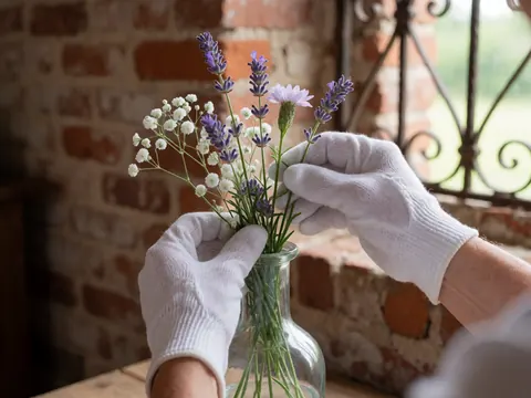Des mains portant des gants blancs placent des fleurs sauvages dans un vase en verre transparent devant un mur de briques rouges et un cadre en fer forgé.
