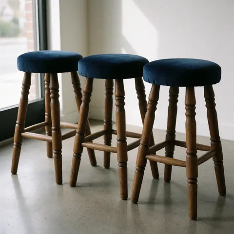 Trois tabourets restaurés dotés d'assises en velours bleu marine et de pieds en bois poli, disposés dans un intérieur baigné de lumière naturelle provenant d'une fenêtre.