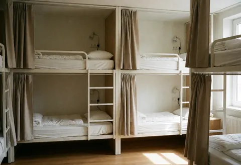 Intérieur d'une chambre d'auberge équipée de lits superposés, rideaux beige, lampes de chevet, prises électriques, sol en bois et lumière naturelle entrant par la fenêtre.