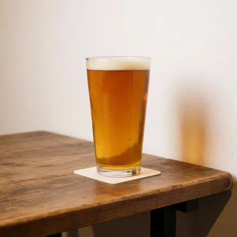 Une bière artisanale servie dans un grand verre avec une mousse blanche, posée sur une table en bois rustique et un sous-verre blanc, devant un mur blanc éclairé de manière chaleureuse.