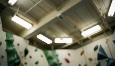 Fond de salle d'escalade avec murs équipés de prises vertes et rouges, plafond aux poutres et lumières fluorescentes flou, idéal pour ajouter du texte.
