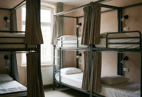 Chambre d'auberge de jeunesse équipée de lits superposés, rideaux, prises électriques et murs en terre cuite. Lumière naturelle entrant par une fenêtre.