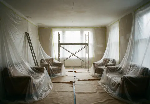 Pièce vide préparée pour les finitions, meubles protégés par du plastique, échafaudages au centre, sol recouvert de papier kraft et lumière naturelle entrant par les fenêtres.