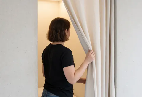 Une femme de dos ferme un rideau blanc dans une chambre d'auberge de jeunesse aux murs en béton et à l'éclairage chaleureux.