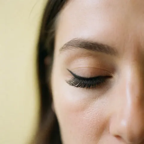 Gros plan d'un oeil portant des cils noirs naturels, contre un mur jaune pâle, éclairage doux mettant en valeur la texture et l'effet papillon.