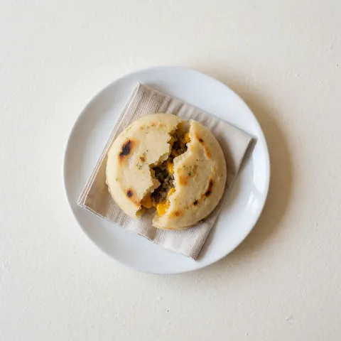 Arepa chaude garnie de fromage fondu et viande, servie sur une assiette blanche posée sur un linge en lin, éclairage clair et net, idéale pour un menu.