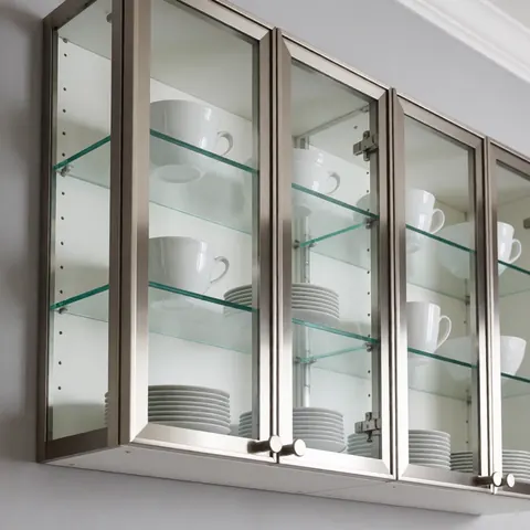 Armoire de cuisine moderne avec portes en verre et cadres en acier inoxydable, exposant assiettes et tasses blanches sur des étagères vitrées.