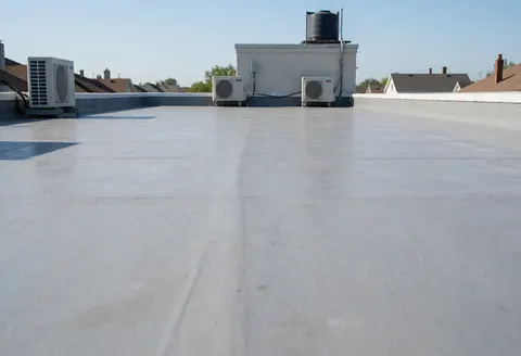 Un toit plat en béton gris préparé pour l'imperméabilisation, avec des unités de climatisation et un réservoir d'eau en arrière-plan sous un ciel bleu clair.