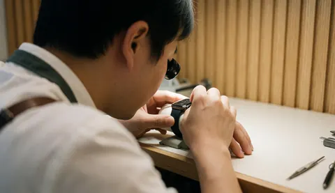 Un horloger examine attentivement un smartwatch avec une loupe dans un atelier en bois clair, entouré d'outils sous une lumière chaleureuse.
