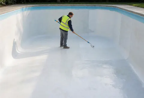 Un ouvrier portant un gilet de sécurité applique un enduit blanc à l'aide d'un long rouleau sur le mur intérieur d'une piscine vide en béton, éclairée par la lumière naturelle.