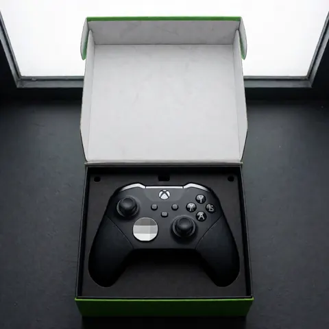 Manette de jeu vidéo noire avec détails argentés, présentée dans sa boîte d'origine ouverte sur une surface grise sombre éclairée par la lumière naturelle.