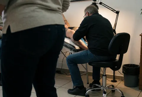 Un tatoueur en lunettes et chemise noire réalise un travail sur la jambe d'un client dans un studio aux sols carrelés, éclairé par une lampe.