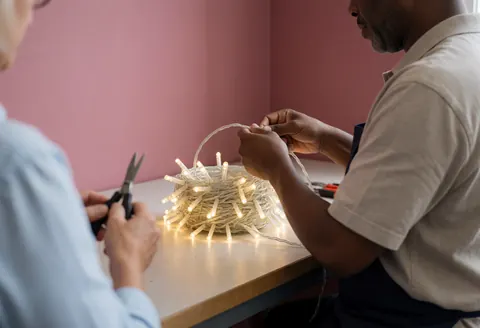 Dans un atelier au mur rose, une personne enroule des câbles lumineux tandis qu'une autre tient des ciseaux, évoquant un environnement de travail pratique et organisé.