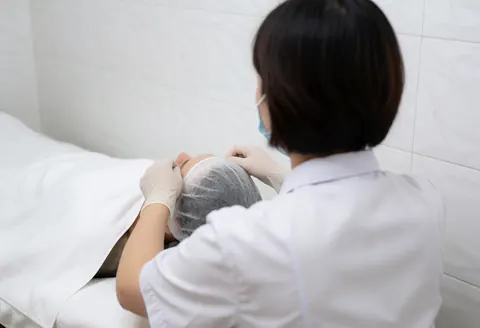 Un dermatologue ajuste un casque de refroidissement sur la tête d'un client allongé sur une table couverte d'une serviette blanche dans une clinique aux murs carrelés.