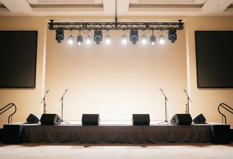 Scène vide équipée de projecteurs, microphones, haut-parleurs et grands écrans sur fond de murs beiges, prête pour des présentations.