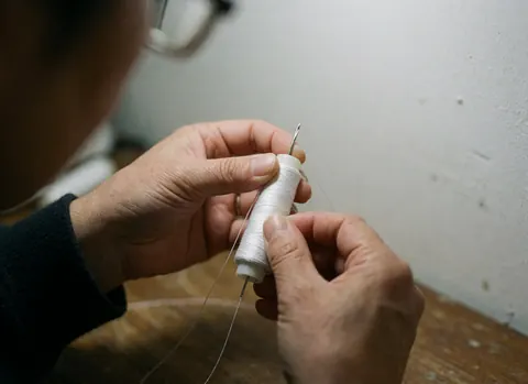 Mains adultes enfilent un fil blanc sur une table en bois avec une aiguille métallique, fond flou.