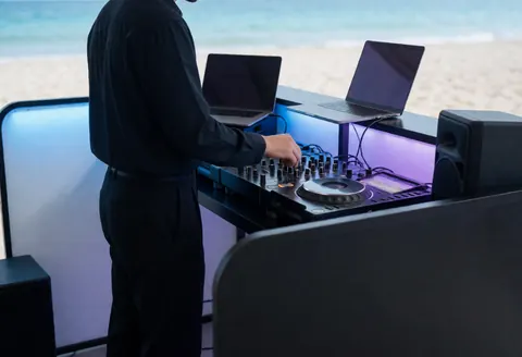 Un dj professionnel ajuste son mixeur dans une cabine de plage équipée de laptops et d'enceintes, avec un arrière-plan flou de la plage et des lumières bleues et violettes.
