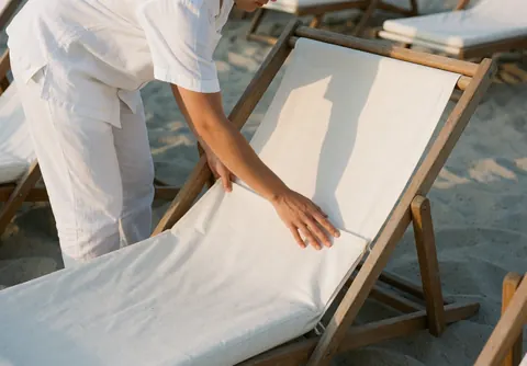 Un travailleur en uniforme blanc ajuste un coussin sur une hamac en bois, sur une plage de sable clair sous un soleil chaud.