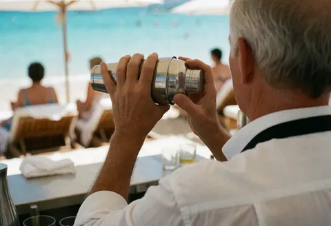 Vue arrière d'un bartender secouant un shaker, mains et outil visibles, fond flou avec eau et chaises.