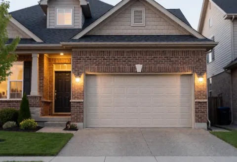 Résidence avec porte de garage beige, façade en brique et enduit, éclairage chaleureux à l'entrée, jardin bien entretenu et rue résidentielle en arrière-plan.
