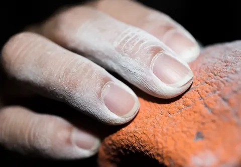 Gros plan d'une main adulte recouverte de poudre blanche agrippant fermement une surface rocheuse orange rugueuse, éclairage direct mettant en valeur la texture.