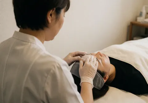 Un thérapeute en gants applique une crème blanche sur le front d'un client allongé. Ambiance calme avec murs blancs et éclairage professionnel.