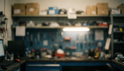 Fond d'atelier avec étagères, outils et un ordinateur au premier plan flou, éclairage chaud aux tons bleus et gris.