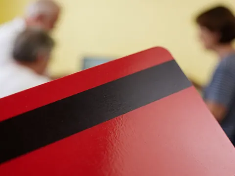 Carte rouge avec bande magnétique noire, fond flou avec personnes devant un mur jaune.